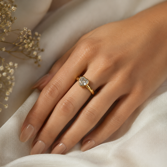 Hand wearing a gold ring with a heart-shaped diamond on a soft fabric background