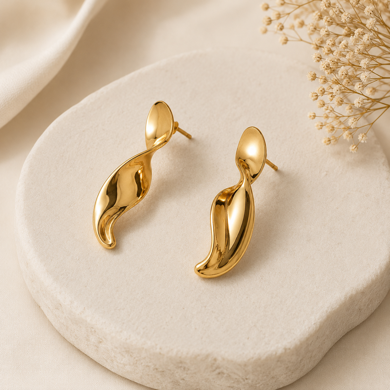 Gold earrings on a beige stone surface with dried flowers.