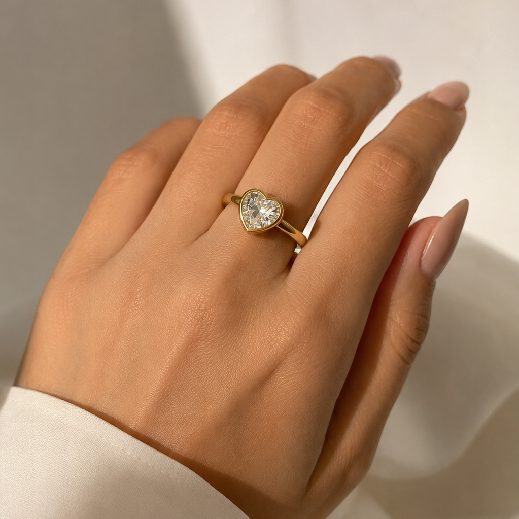 A gold ring with a heart-shaped clear gemstone set on a person's ring finger, shown against a soft white background.