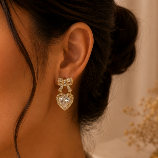 Close-up of an ear wearing a gold bow-shaped earring with a heart detail, against a blurred background.