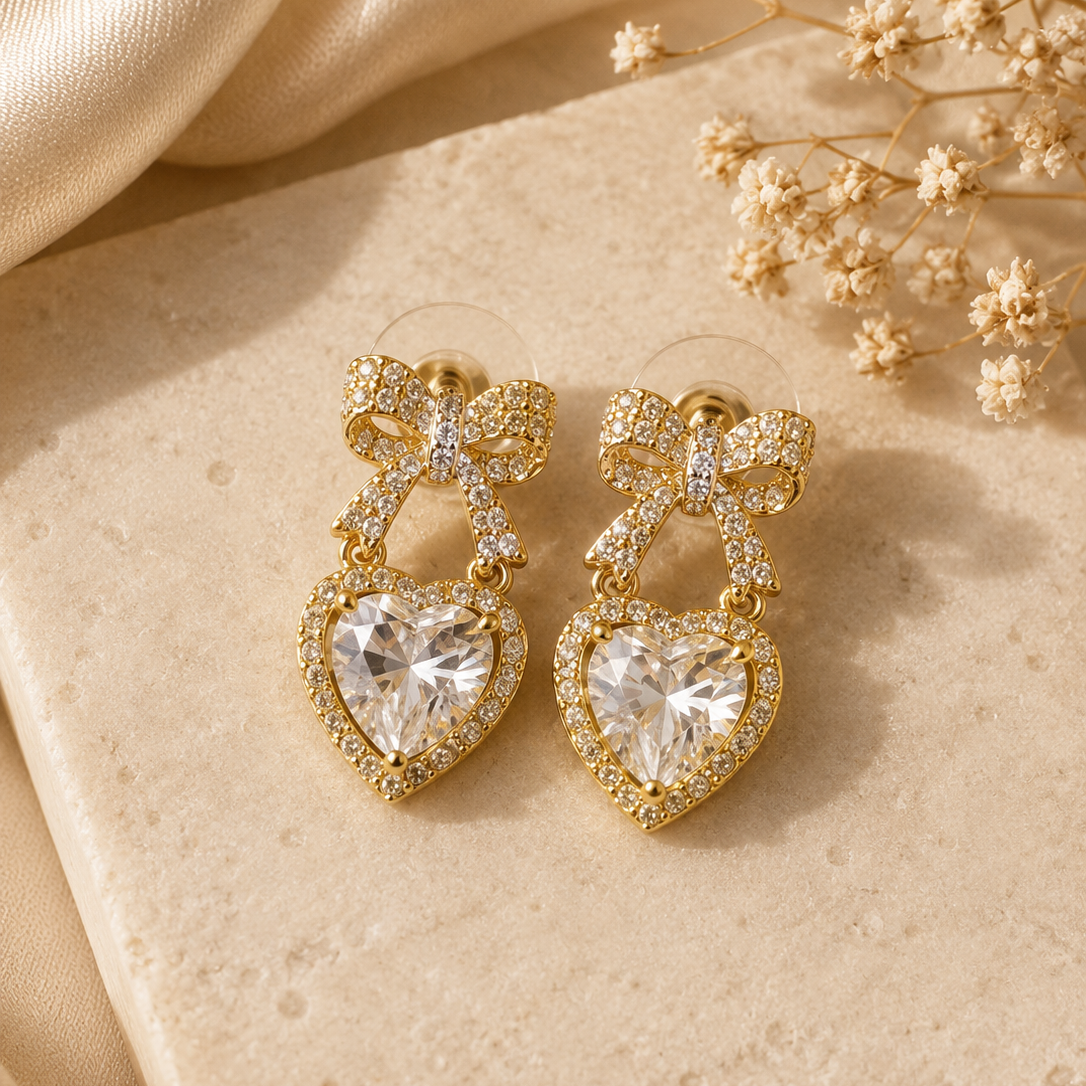 Gold bow-shaped earrings with heart details on a beige surface with dried flowers.