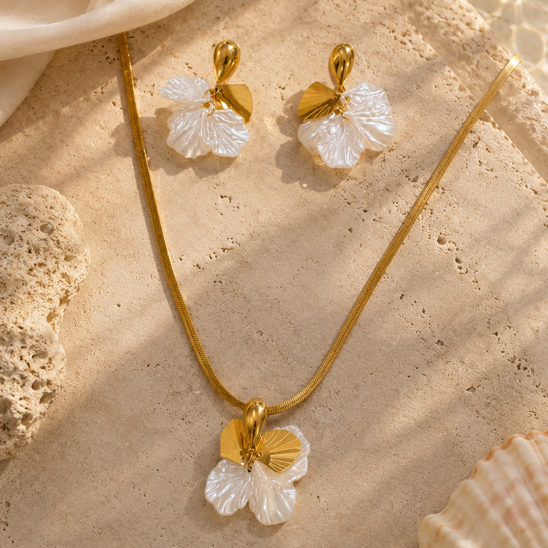 Gold necklace and earrings with white floral pendants on a beige stone surface.