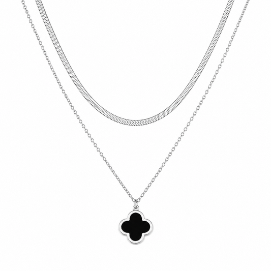 A silver-toned double-strand necklace featuring a textured chain and a delicate cable chain with a black clover pendant, shown on a plain white background.
