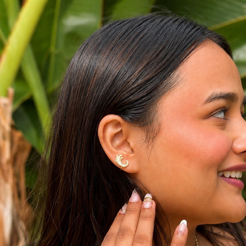 A woman wearing a gold necklace with a small shell pendant, a gold bangle, and a gold ring, shown in a close-up profile view against a background of green tropical leaves.
