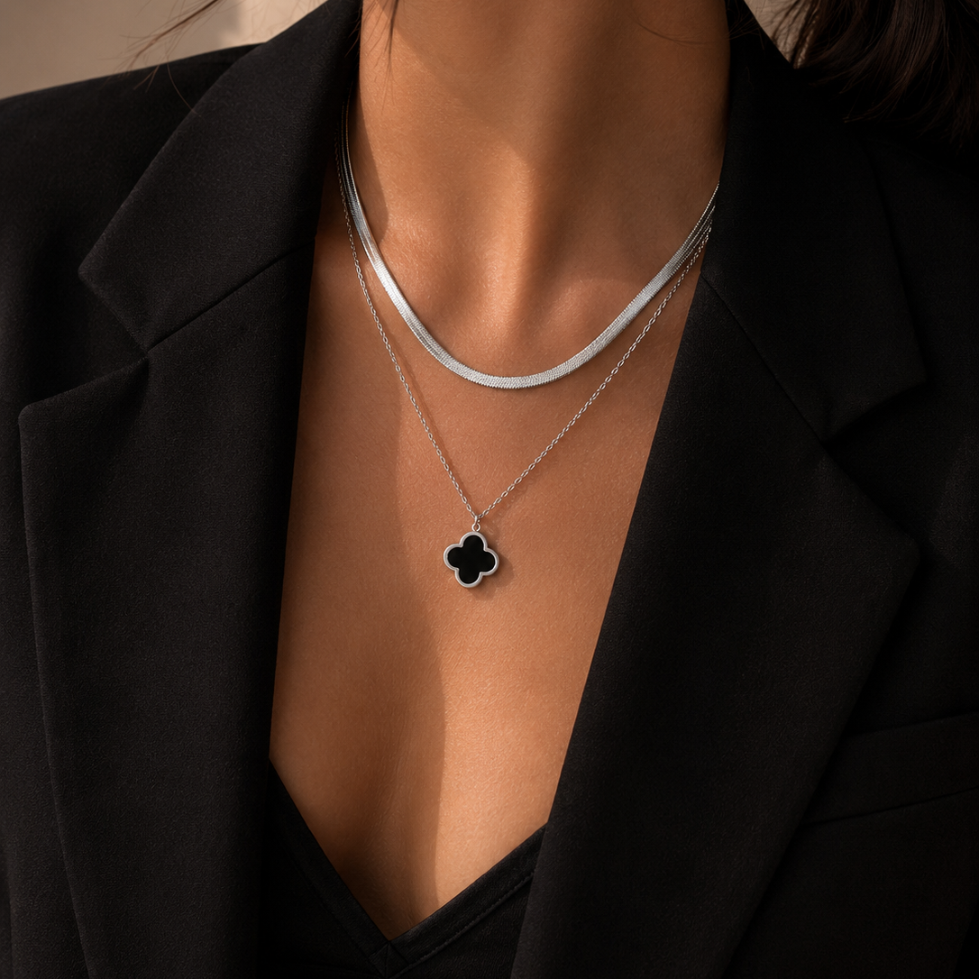 A close-up of a model wearing a layered silver necklace set, featuring a thin chain and a delicate chain with a black clover pendant, styled with a black blazer.