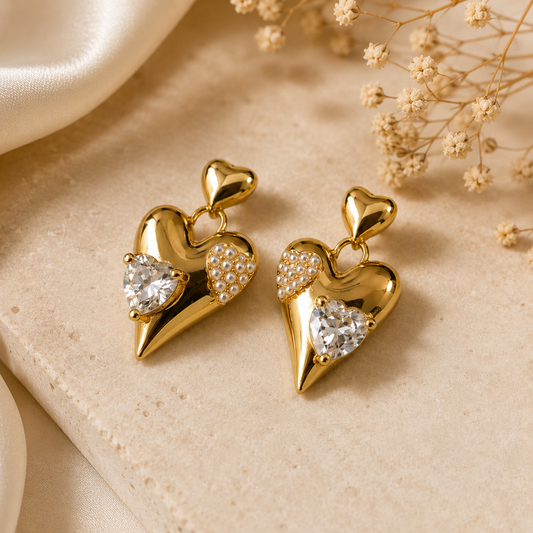 Gold heart-shaped earrings with gemstones on a beige surface with dried flowers.
