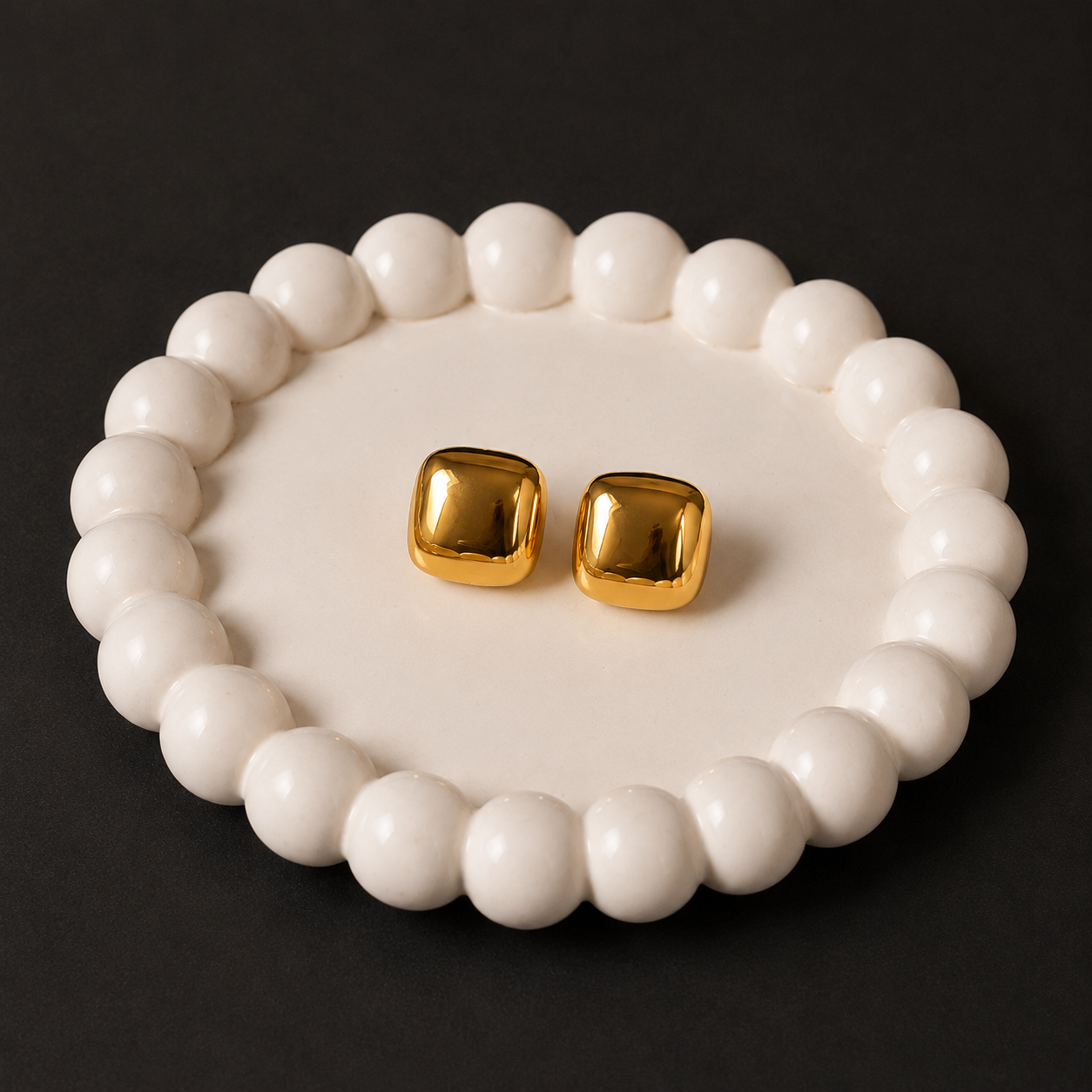 Gold square earrings on a white beaded dish against a black background