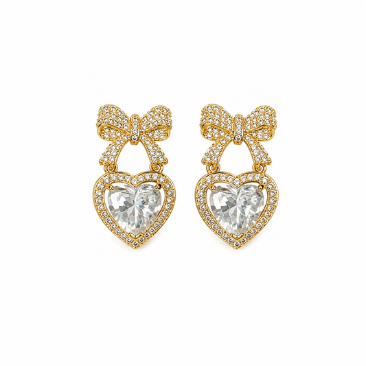 A pair of gold heart-shaped drop earrings featuring a pavé-set clear heart gemstone and a matching pavé bow above it, shown on a plain white background.