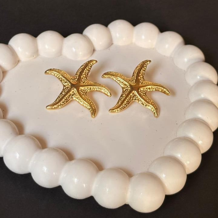 Gold starfish earrings on a white beaded bracelet against a dark background