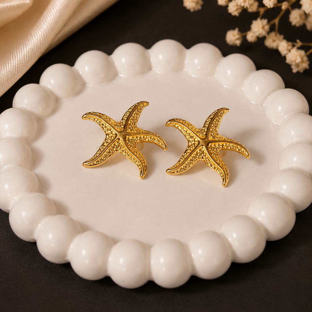 Gold starfish-shaped earrings on a white beaded dish with a dark background